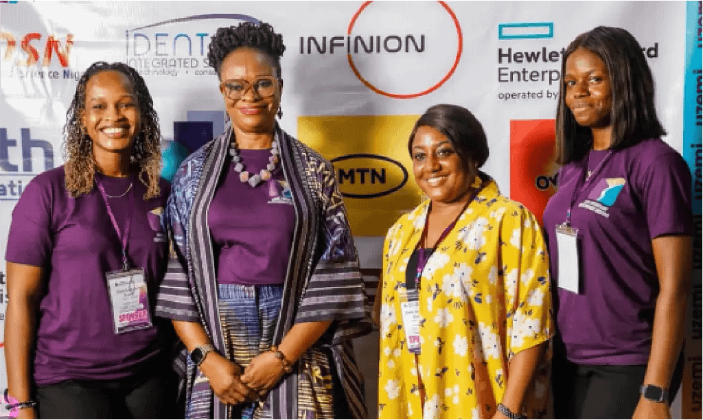 Four women stand together at a conference or expo, smiling at the camera; three wear purple shirts with name badges, one wears a yellow floral dress, banner with sponsor logos behind them.