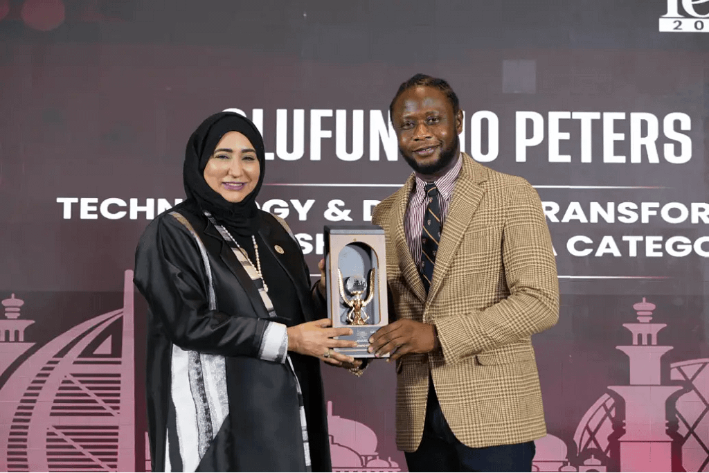 Two well-dressed individuals on a stage pose with a trophy, as the man hands it to the woman wearing a hijab and a black outfit.