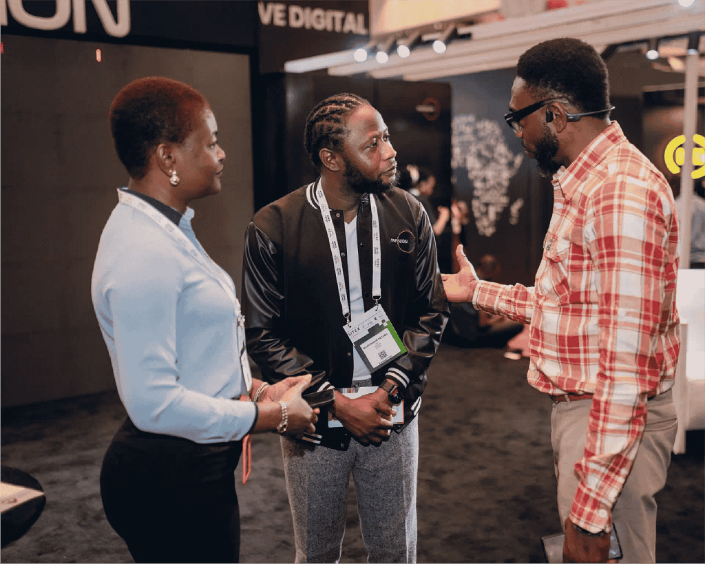 Three adults wearing conference badges converse in an exhibit hall at a busy event, center man with braided hair in a black jacket flanked by a woman and a man in a plaid shirt.